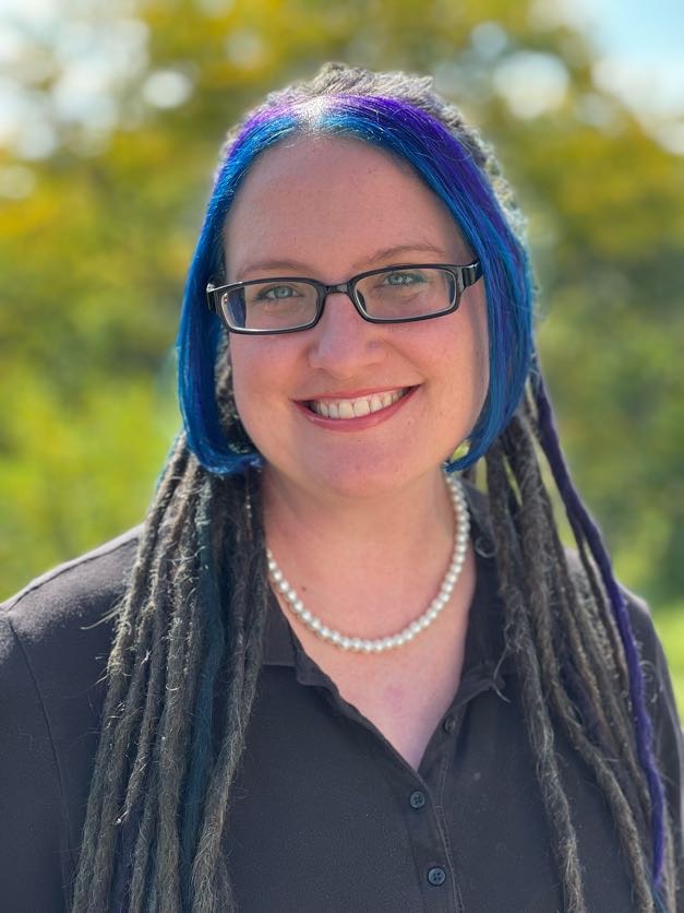 Rose Howell smiling outdoors with long dark hair, wearing glasses, a black shirt, and a necklace, with a softly blurred green background