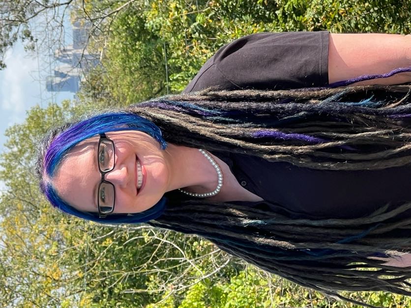 Rose Howell smiling outdoors with long dark hair, glasses, a black shirt, and a necklace, with greenery in the background and a city skyline visible in the distance